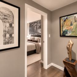 Hallway with gray walls, a large abstract black-and-white print on the left, a colorful painting on the right, and a sculptural wooden statue by a pedestal, leading to a bedroom.