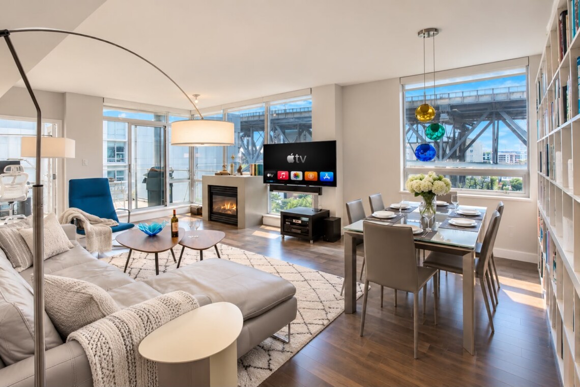 Open-concept living and dining area in a bright apartment: gray sofa, blue chair, fireplace, and a TV near the windowed wall; bookshelf on the right.