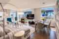 Open-concept living and dining area in a bright apartment: gray sofa, blue chair, fireplace, and a TV near the windowed wall; bookshelf on the right.