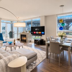 Open-concept living and dining area in a bright apartment: gray sofa, blue chair, fireplace, and a TV near the windowed wall; bookshelf on the right.