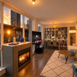 Sunlit city apartment living room with large windows, a fireplace, TV, and a tall bookshelf surrounding a dining area.