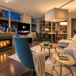 Contemporary living room at dusk with a lit fireplace, large windows showing city buildings, a TV stand, and a blue accent chair near a gray sectional.