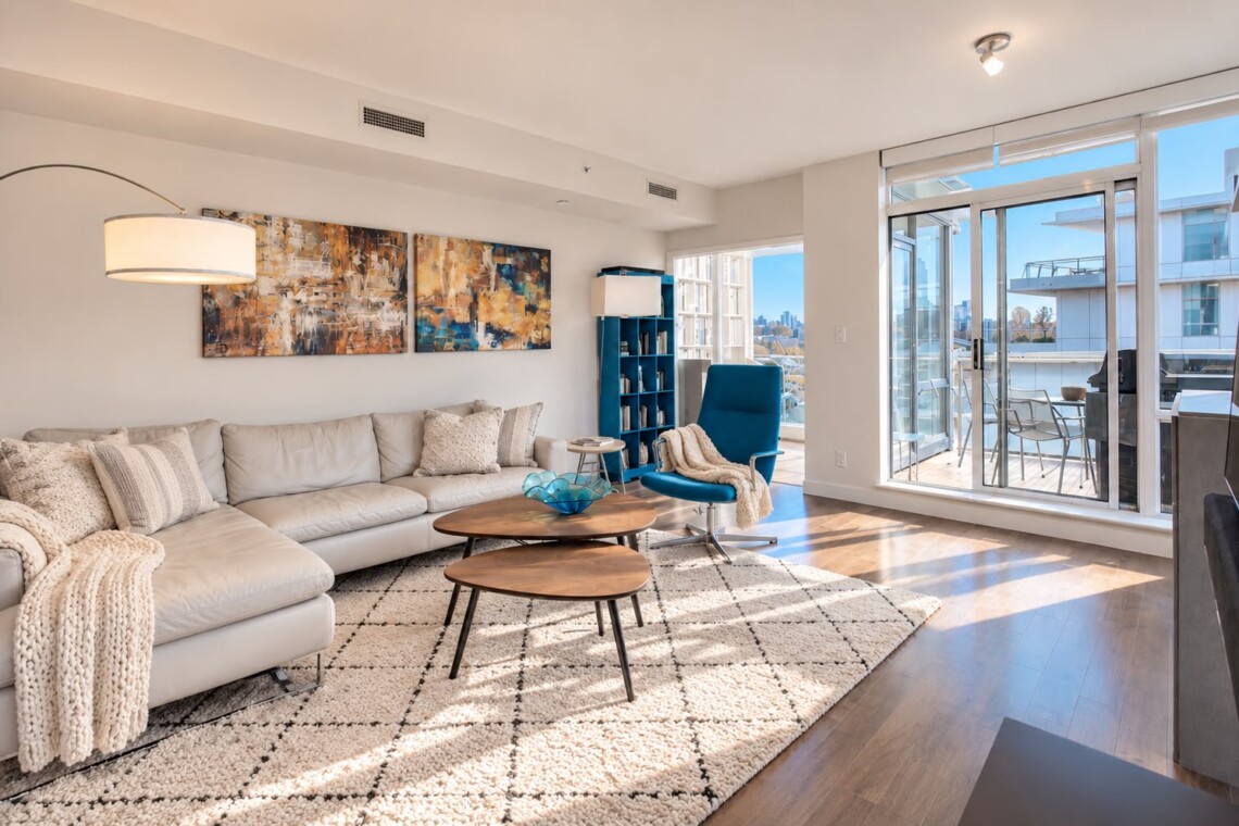 Modern living room with a light gray sectional, cream throws, abstract art, and a blue recliner beside a large windowed balcony.