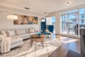 Modern living room with a light gray sectional, cream throws, abstract art, and a blue recliner beside a large windowed balcony.