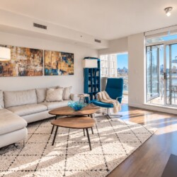Modern living room with a light gray sectional, cream throws, abstract art, and a blue recliner beside a large windowed balcony.