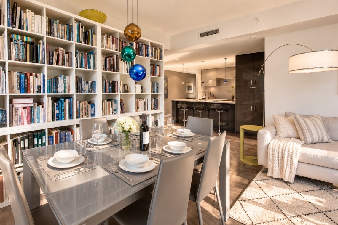 Open-concept living and dining area with a tall white bookshelf wall, a glass dining table set for six, and a beige sofa nearby.