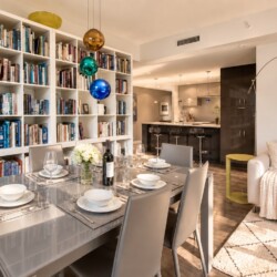 Open-concept living and dining area with a tall white bookshelf wall, a glass dining table set for six, and a beige sofa nearby.
