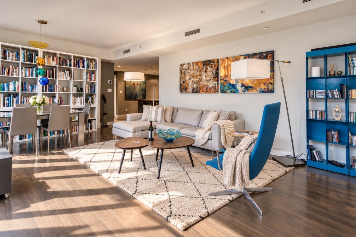 Bright living room with a gray sectional, blue accent chair, two round wooden coffee tables, and a cream rug under soft lighting.