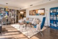Bright living room with a gray sectional, blue accent chair, two round wooden coffee tables, and a cream rug under soft lighting.