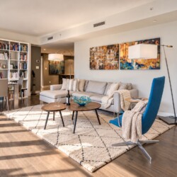 Bright living room with a gray sectional, blue accent chair, two round wooden coffee tables, and a cream rug under soft lighting.