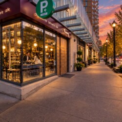 Cafe storefront at dusk with warm interior lights, two people dining inside, on a city sidewalk lined with lamps and trees.