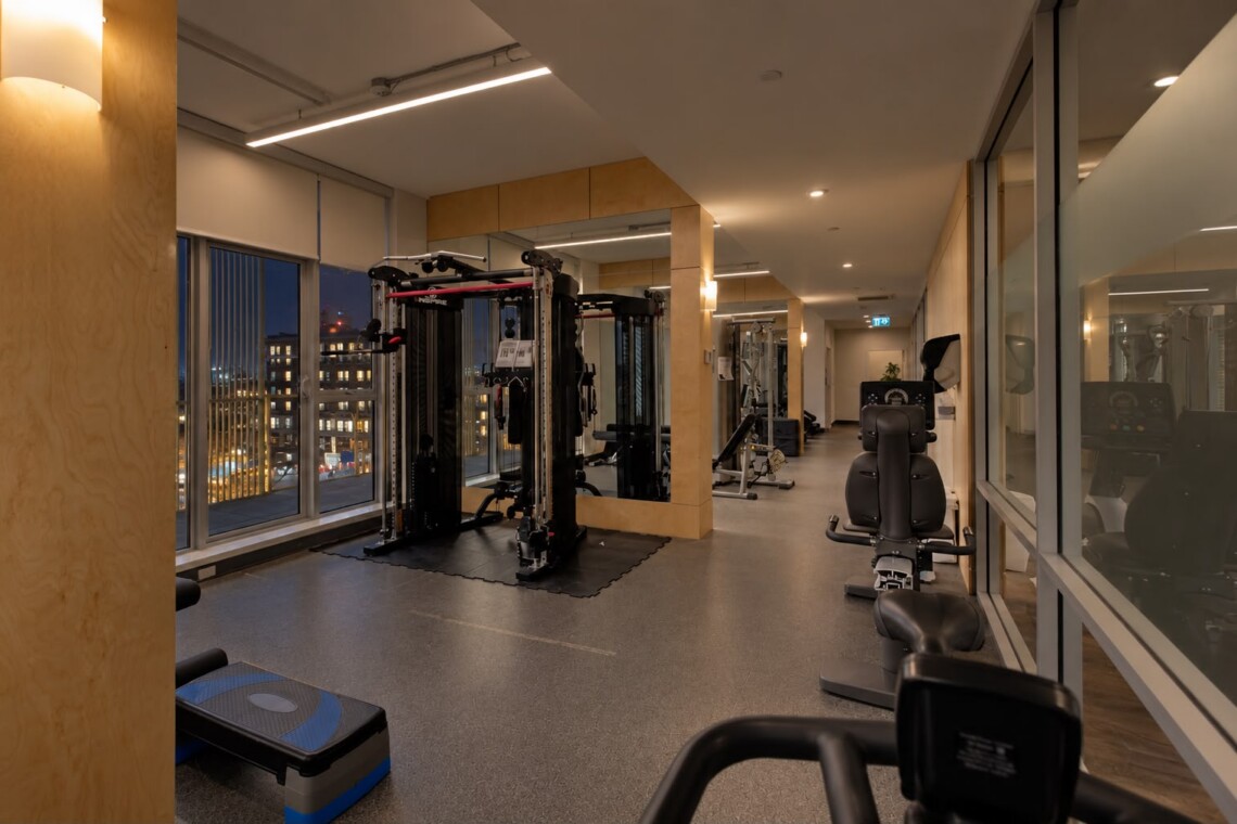 Modern gym interior at night with cable machines, weight racks, and mats, seen through large windows showing city lights outside.