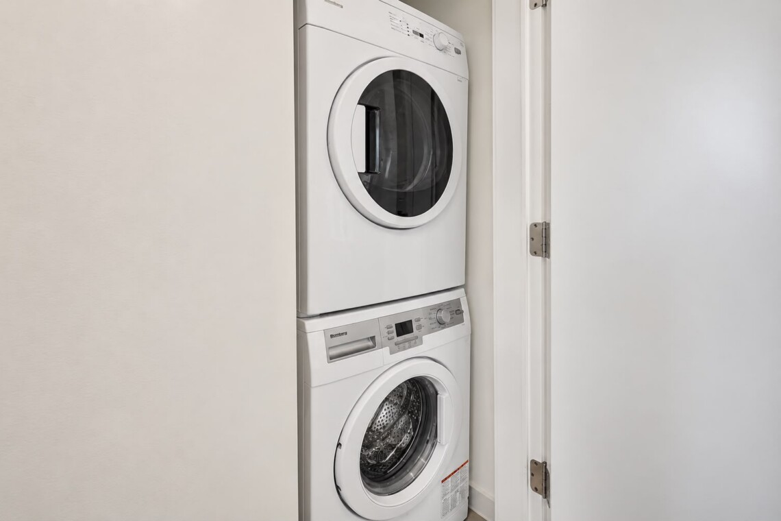 Stacked white front-loading washer and dryer in a narrow laundry closet with a white door open