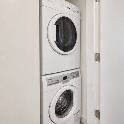 Stacked white front-loading washer and dryer in a narrow laundry closet with a white door open