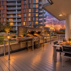 Outdoor rooftop patio at dusk with a long gas grill setup and a dining table set for six, overlooking modern apartment buildings.