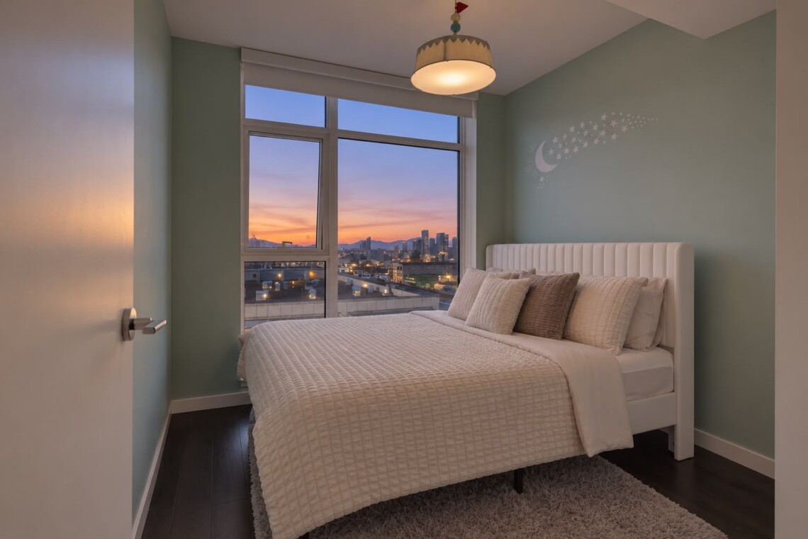 Cozy bedroom with a white upholstered bed, pastel green walls, and a moon/star wall decal overlooking a city sunset from a large window.