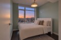 Cozy bedroom with a white upholstered bed, pastel green walls, and a moon/star wall decal overlooking a city sunset from a large window.