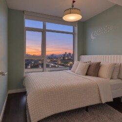 Cozy bedroom with a white upholstered bed, pastel green walls, and a moon/star wall decal overlooking a city sunset from a large window.