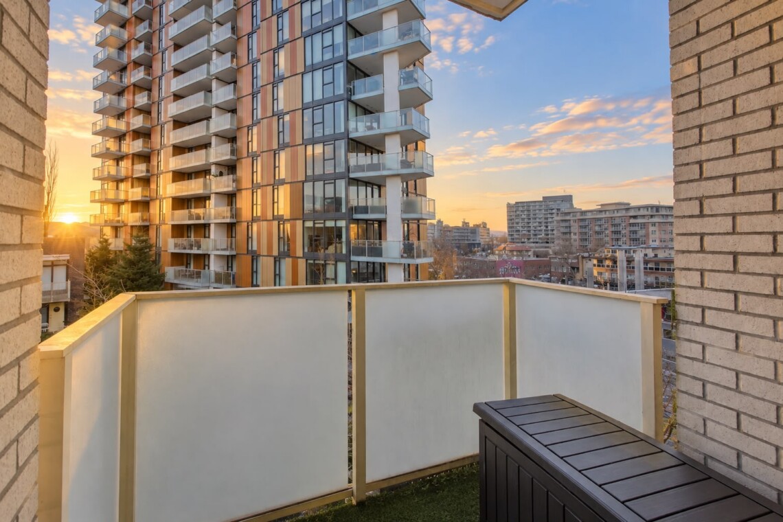 Sunset cityscape viewed from a balcony with a glass-paneled railing and a neighboring modern apartment building nearby.
