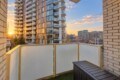 Sunset cityscape viewed from a balcony with a glass-paneled railing and a neighboring modern apartment building nearby.