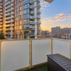 Sunset cityscape viewed from a balcony with a glass-paneled railing and a neighboring modern apartment building nearby.