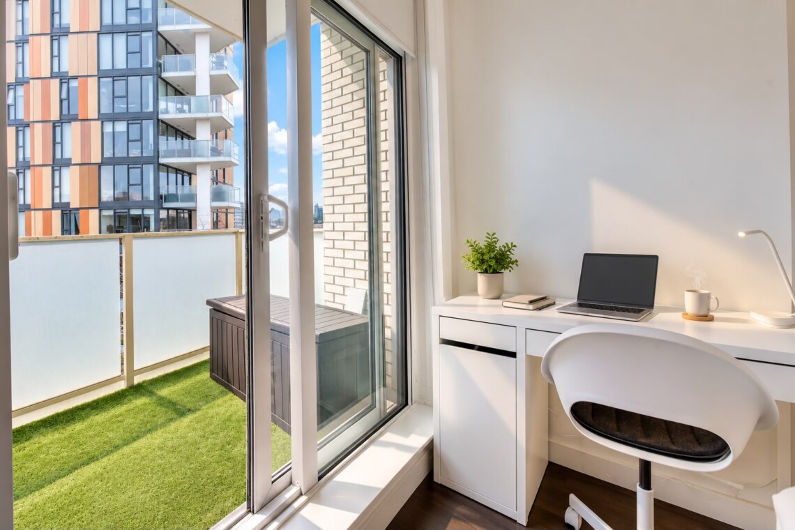 Bright home office with a white desk by a glass balcony door, featuring a laptop, plant, books, and a coffee mug.