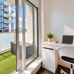 Bright home office with a white desk by a glass balcony door, featuring a laptop, plant, books, and a coffee mug.
