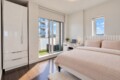 Bright modern bedroom with white furniture, pink bedding, and a balcony sliding door showing a city view.