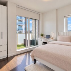 Bright modern bedroom with white furniture, pink bedding, and a balcony sliding door showing a city view.