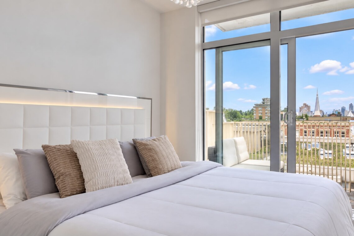 Bright hotel-style bedroom with white tufted headboard, beige pillows, and a balcony door overlooking a city skyline.