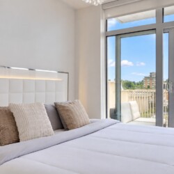 Bright hotel-style bedroom with white tufted headboard, beige pillows, and a balcony door overlooking a city skyline.