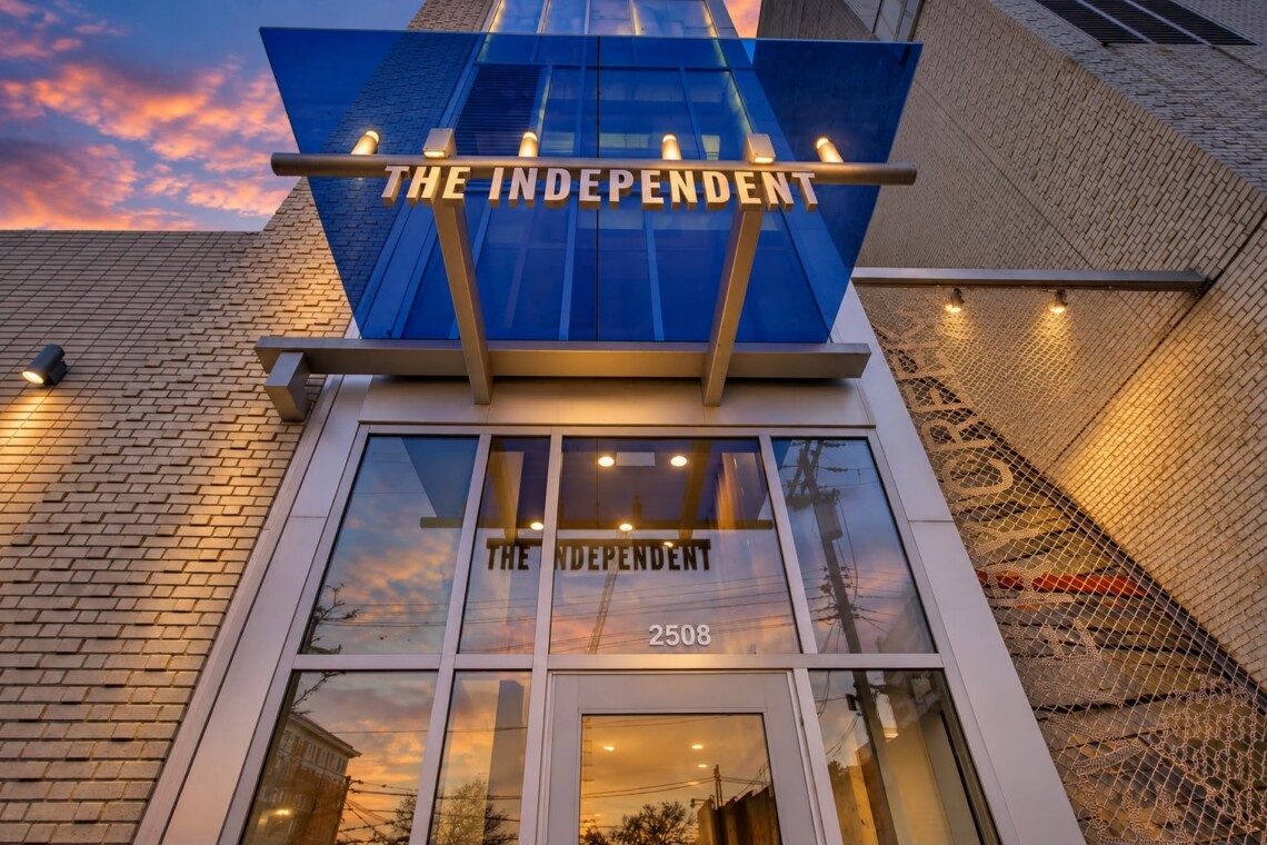 Facade of a modern building with a blue glass canopy and 'THE INDEPENDENT' sign over the entrance, sunset reflecting in glass, 2508 address visible
