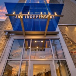 Facade of a modern building with a blue glass canopy and 'THE INDEPENDENT' sign over the entrance, sunset reflecting in glass, 2508 address visible