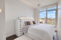 Bright modern bedroom with a white tufted headboard, gray bedding, and a tall white dresser; large window opens to a balcony with a city view.