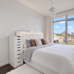 Bright modern bedroom with a white tufted headboard, gray bedding, and a tall white dresser; large window opens to a balcony with a city view.