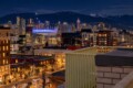 Nighttime city skyline with illuminated skyscrapers, a blue-lit stadium, and snow-capped mountains in the distance behind the city.