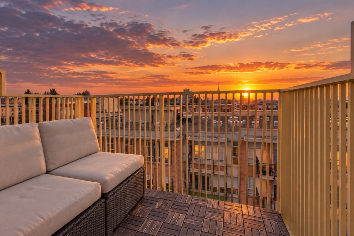 Rattan outdoor sofa with beige cushions on a wooden deck, overlooking a city at sunset with a yellow railing.