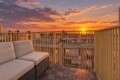 Rattan outdoor sofa with beige cushions on a wooden deck, overlooking a city at sunset with a yellow railing.