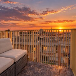 Rattan outdoor sofa with beige cushions on a wooden deck, overlooking a city at sunset with a yellow railing.