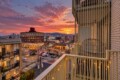Sunset over a downtown street viewed from a balcony, with warm orange clouds, city buildings, and distant mountains on the horizon.
