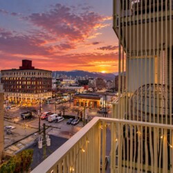Sunset over a downtown street viewed from a balcony, with warm orange clouds, city buildings, and distant mountains on the horizon.