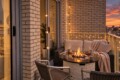 Cozy balcony seating area at sunset with a fire pit, wicker sofa, string lights, and a warm glow over a city skyline.