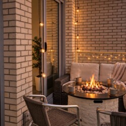 Cozy balcony seating area at sunset with a fire pit, wicker sofa, string lights, and a warm glow over a city skyline.