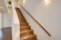 Wooden staircase with a dark wood handrail along a white wall, a potted plant on the landing, and warm wall lights in a modern interior.