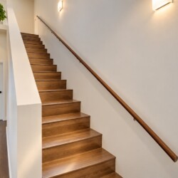 Wooden staircase with a dark wood handrail along a white wall, a potted plant on the landing, and warm wall lights in a modern interior.