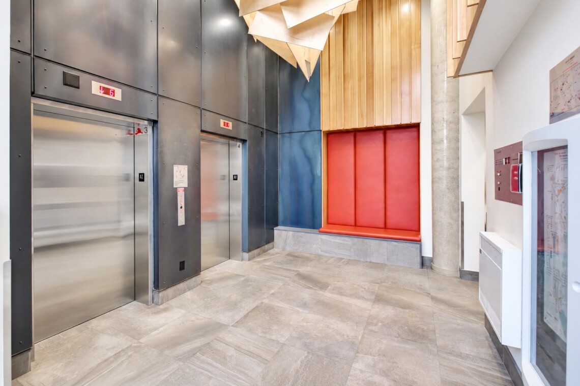 Modern elevator lobby with two stainless steel doors, blue wall panels, and a red cushioned bench against a wood-paneled wall.
