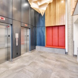 Modern elevator lobby with two stainless steel doors, blue wall panels, and a red cushioned bench against a wood-paneled wall.