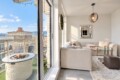 Bright living room with beige sectional facing a glass door to a city-view balcony; a circular fire pit on a white stone table outside.