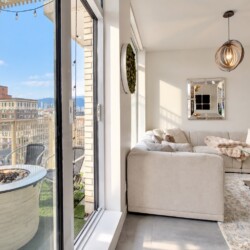 Bright living room with beige sectional facing a glass door to a city-view balcony; a circular fire pit on a white stone table outside.