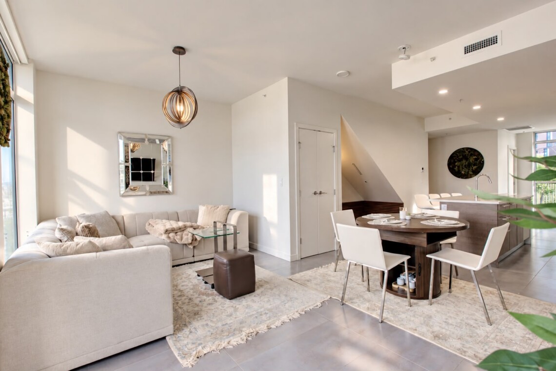 Open-plan living and dining area with a beige sectional sofa, glass coffee table, and a round dining table with white chairs near a kitchen island.